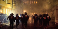Mehrere Polizisten laufen nachts durch eine Straße, Feuerwerk beleuchtet die Szene