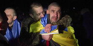 Ein ukrainischer Kriegsgefangener mit einer Flagge der Ukraine um die Schultern wird von einem Mann umarmt und ist sichtlich bewegt