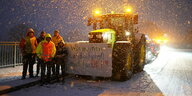 Fünf Menschen in Warnwesten stehen im Schneegestöber nachts neben einem Traktor, an dem ein Transparenz befestigt ist: "Wir machen Landwirtschaft auch für Dich!"