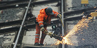 Ein Arbeiter in orangener Arbeitskleidung werkelt an Gleisen. Funken sprühen.