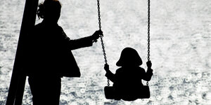 Schattenriss - Frau mit Kind auf dem Spielplatz, das Kind schaukelt, die Mutter gibt der Schaukel einen Schubs