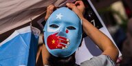Die Maske eines gesichts ist in den Farben der Uiguren bemalt, eine Hand, die in den Nationalfarben von China bemalt ist, verschliesst den Mund