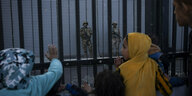 Kinder stehen vor einem Grenzzaun. Hinter dem Zaun stehen bewaffnete Soldaten.