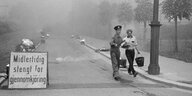Schwarz-Weiß-Aufnahme 60er Jahre, ein Uniformierter und ein Mann mit Eimer am Straßenrand, auf der Straße viele kleine Brände