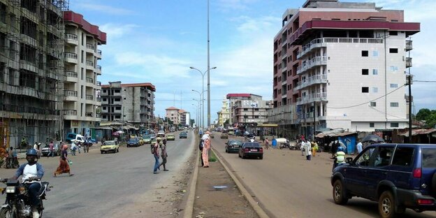 Straße in Conakry. Rechts und links Häuser und Bauruinen. In der MItte ein schmaler Grünstreifen mit Straßenlaternen.