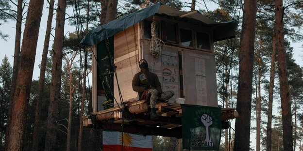 Eine Person sitzt auf den Planken eines Baumhauses im Wald