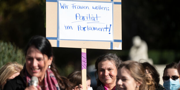 Drei Teilnehmerinnen einer Kundgebung stehen neben einem Plakat mit der Aufschrift „Wir Frauen wollen Parität im Parlament!“, eine hält ein Mirkofon