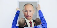 Zwei Arbeiter in blauer Arbeitskleidung hängen ein Porträt des russischen Präsidenten Wladimir Putin auf