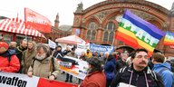 Teilnehmende beim Ostermarsch 2017 mit Plakaten und Fahnen vor dem Bremer Hauptbahnhof. Zu sehen sind unter anderem eine "Peace"-Flagge in Regenbogenfarben, ein Transparent mit der Friedenstaube, sowie ein Spruchband der DKP und eine Fahne der Partei Die