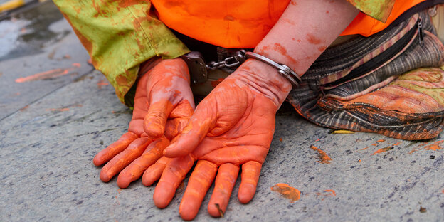 Zwei in Handschellen gefesselte und mit orangener Farbe beschmierte Hände auf Asphalt