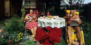 Ein älteres Ehepaar sitzt im sommerlichen Garten an einem gedeckten Tisch. Der Mann liest Zeitung, sie ein Magazin zum Thema Wohnen