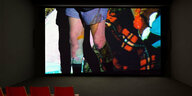 Im Videoraum des KINDL-Zentrums ist ein Video auf die Leinwand projiziert. Die Beine einer Person sind zu sehen, sie trägt Boxershorts, Strumpfbänder und schwarze Socken. Hinter den beiden ist ein Stoff mit Schottenmuster zu sehen. Im vorderen Bildbereich sind orangene Stuhllehnen der Raumbestuhlung zu erkennen.