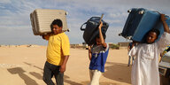 Drei Personen laufen mit Koffern auf ihren Schultern durch eine wüstenähnliche Landschaft
