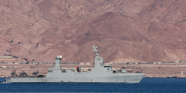 Ein graues Kriegsschiff fährt auf Wasser, im Hintergrund eine sandige Landschaft
