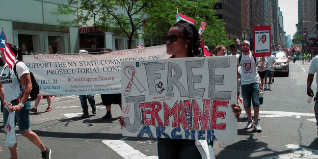Aufnahme einer Demonstration. Eine Afroamerikanerin hält ein großes Transparent in den Händen, auf dem steht "Free Jermaine Archer".