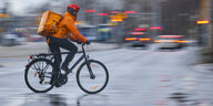 Ein Fahrradfahrer im orangen Lieferando Outfit mit Fahrradhelm und Lieferrucksack fährt auf regennasser Straße über eine Fahrbahn