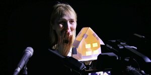 Das Gesicht einer lächelnden Frau hinter einem kleinen Modellhaus, das von innen leuchtet