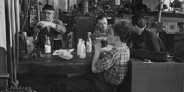 Schwarz-Weiß-Aufnahme: Vier Männer sitzen in einer Werkhalle an einem Tisch