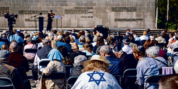 Teilnehmende einer Gedenkveranstaltung sitzen auf Klappstühlen auf dem Gelände der Gedenkstätte Bergen-Belsen.