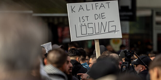 Mehrere Demonstranten stehen beieinander, einer hält ein Schild hoch, auf dem steht "Kalifat ist die Lösung"