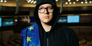 Ein Mann mit Kaputze im Europaparlament mit Europaflagge über der Schulte