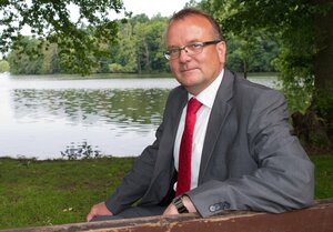 Portrait von Detlef Tabbert - er sitzt in Anzug und roter Krawatte auf einer Bank an einem Badesee