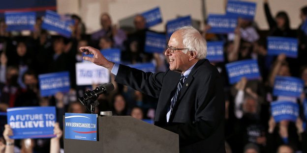 Bernie Sanders am Redpult, im Hintergrund viele Menschen mit blauen Schildern, er lächelt und hebt die Hand.
