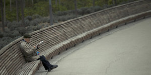 Ein älterer Mann sitzt mit Jacke und Mütze alleine auf der Bank und liest Zeitung.