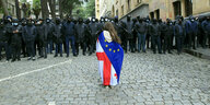 Eine Frau mit wallendem Haar ist in eine Georgische-EU-Fahne gehüllt und steht einer Polizeikette gegenüber