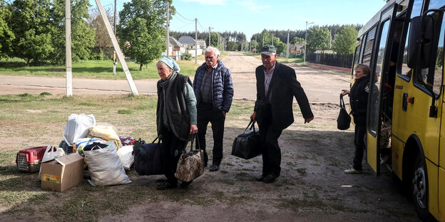 Drei ältere Menschen, die mit viel Gepäck neben einem Bus an einer Haltestelle auf dem Land stehen