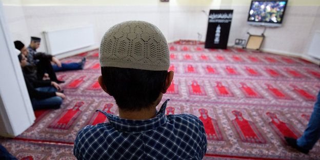Ein Junge mit einer weißen Mütze steht in einem mit Teppich ausgelegten Gebetsraum