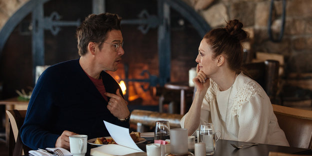Ein Ethan Hawke und eine Julianne Moore sitzen sich in an einem Tisch beim Essen gegenüber, hinter ihnen lodert ein Feuer im Kamin.