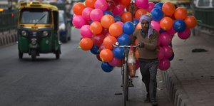 Ein Mann schiebt ein Fahrrad an dem viele bunte Luftballons hängen, eine Straße entlang.