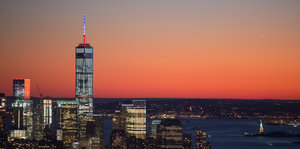 Hinter der Skyline Manhattans mit erleuchteten Fenster zeigt sich ein rosiger Sonnenuntergang.