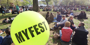 Ein gelber Luftballon, auf dem „Myfest“ steht, dahinter sitzen Menschen auf einer Wiese in der Sonne