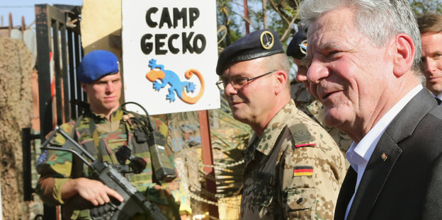 Herr Gauck steht neben zwei Bundeswehrsoldaten und dem Schild „Camp Gecko“.