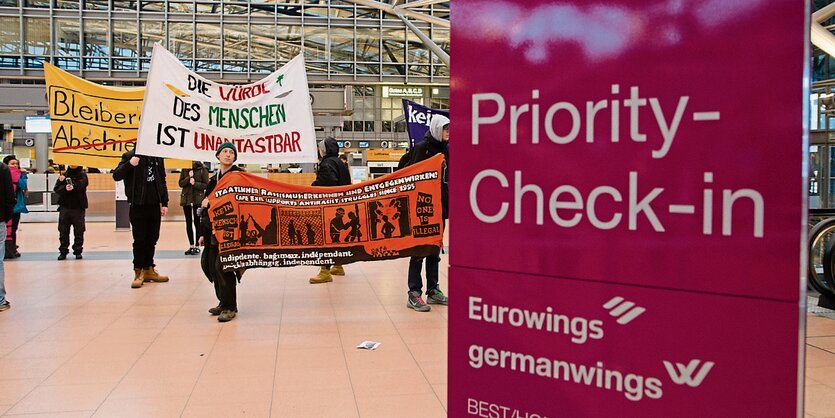 Ein violettes Priority-Check-In-Schild, daneben DemonstrantInnen