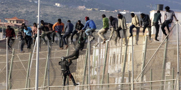 18 Menschen hängen auf einem hohen Zaun. Einer wird von einem Grenzpolizisten heruntergezogen.
