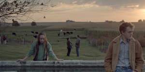 Ein Mädchen und ein junger Mann am Rande einer weiten Landschaft mit Pferden und Abendhimmel.