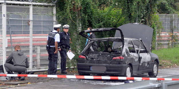 Zwei Polizisten, eine Frau und ein Mann im Overall stehen an einem ausgebrannten Autowrack. Im Vordergrund ist ein Absperrband, im Hintergrund ein Zaun.