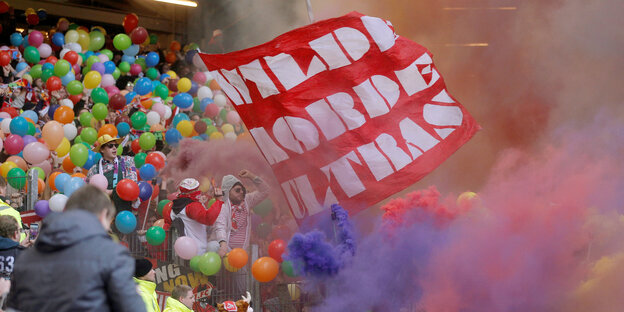 Fußballfans schwingen eine Fahne, halten Luftballons und zünden Bengalos