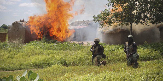 Zwei Soldaten laufen durch hohes Gras. Im Hintergrund brennen Häuser