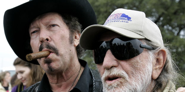 ein Mann mit dicker Zigarre im Mund und Cowboyhut, daneben ein Mann mit Basecap und großer Sonnenbrille