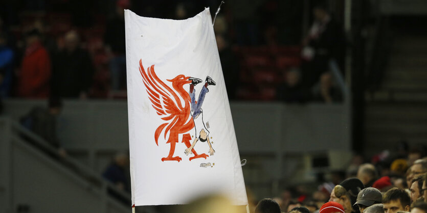 Ein Doppelhalter-Plakat in einem Fußballstadion: Darauf ist ein großer roter Vogel, der eine Person kopfüber im Schnabel hat. Die Person verliert Geld.