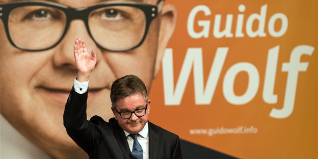 Guido Wolf winkt vor seinem Wahlplakat dem Publikum zu.