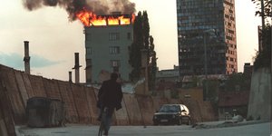 Ein Fahrradfahrer auf einer leeren Straße, im Hintergrund ein brennendes Hochhaus