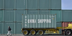 Ein Container mit der Aufschrift „China Shipping“ steht vor vielen aufeinenadre getürmten weiteren Containern. Davor läuft eine Person