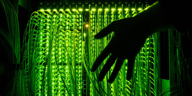 eine Hand vor einem Verteilerpunkt, in dem grün leuchtende Glasfaserkabel zusammenlaufen