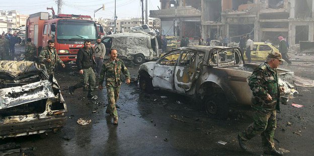 eine Straße mit zerstörten und ausgebrannte Autos, Menschen in Uniform, im Hintergrund ein komplett zerstörtes Gebäude