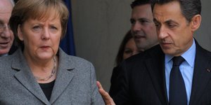 Sarkozy legt seine Hand an Merkels Arm. Beide schauen grimmig.